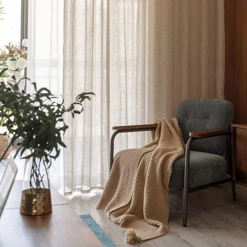 Fauteuil confortable avec plaid dans un salon lumineux, idéal pour la détente. Rideaux élégants laissant passer la lumière naturelle, plante d'intérieur et décoration moderne pour un espace de vie accueillant et chaleureux.