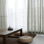 Rideaux élégants pour salon moderne, apportant une touche de luminosité et de douceur naturelle. Ambiance zen avec table basse en bois et coussins de sol tressés, créant un espace chaleureux et accueillant. Parfait pour une décoration intérieure raffinée et minimaliste.
