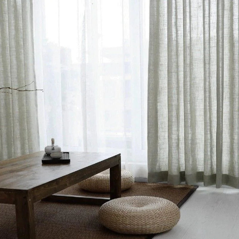 Rideaux élégants pour salon moderne, apportant une touche de luminosité et de douceur naturelle. Ambiance zen avec table basse en bois et coussins de sol tressés, créant un espace chaleureux et accueillant. Parfait pour une décoration intérieure raffinée et minimaliste.