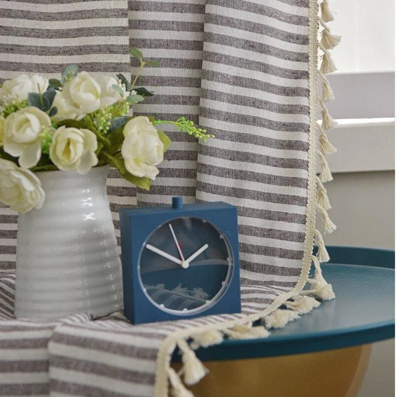 Horloge carrée minimaliste bleue posée sur une table bleue, accompagnée d'un vase blanc avec des fleurs blanches. Décoration élégante et moderne, idéale pour un intérieur contemporain. Rideau à rayures et franges en arrière-plan pour une ambiance tendance et raffinée.