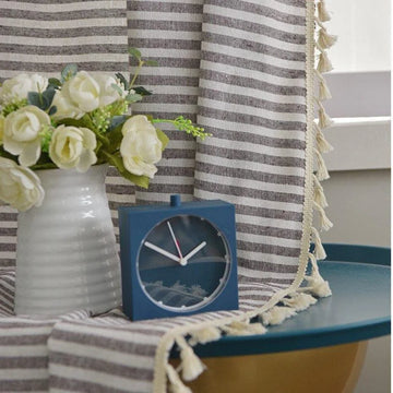 Horloge carrée minimaliste bleue posée sur une table bleue, accompagnée d'un vase blanc avec des fleurs blanches. Décoration élégante et moderne, idéale pour un intérieur contemporain. Rideau à rayures et franges en arrière-plan pour une ambiance tendance et raffinée.