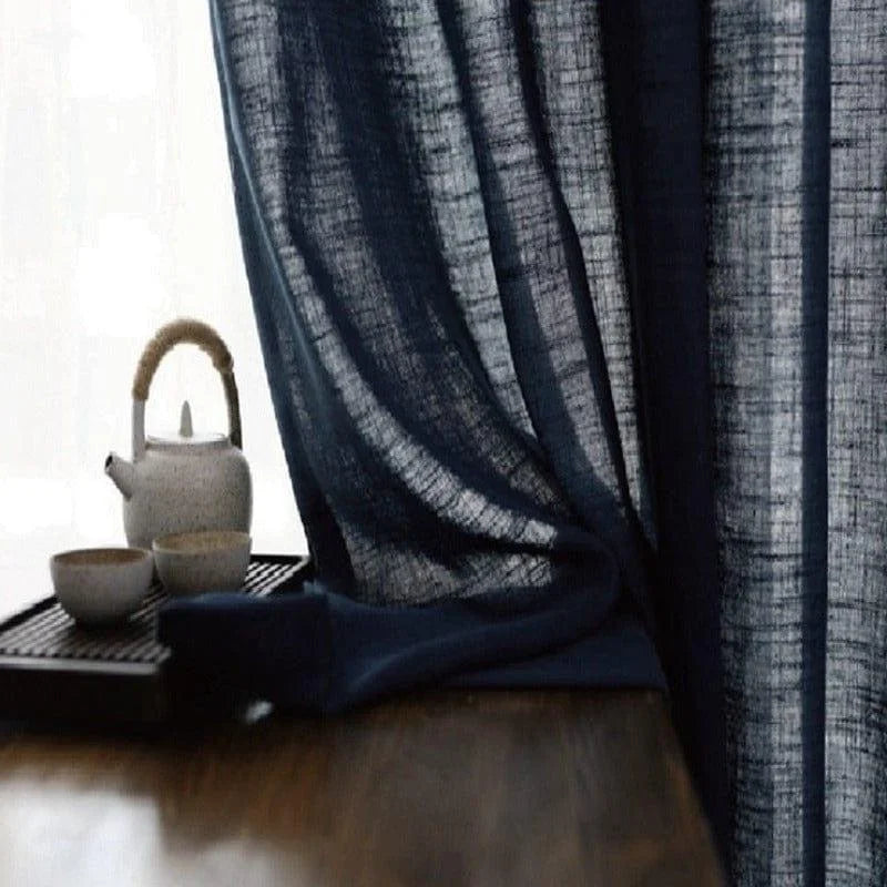 Rideau bleu foncé drapé élégamment, laissant filtrer la lumière naturelle, posé à côté d'une table en bois sur laquelle repose un ensemble à thé en céramique rustique. Cet agencement crée une atmosphère paisible et raffinée dans votre espace de vie.