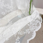 Rideau blanc avec motif floral en transparence sur une table en extérieur, créant une atmosphère élégante et lumineuse. Accessoire de décoration idéal pour ajouter une touche de sophistication à votre intérieur. Parfait pour les salons ou chambres, ce rideau combine esthétique et fonctionnalité.