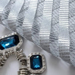 Broderie argentée élégante avec ornements bleus. Conception raffinée idéale pour décorer des vêtements ou accessoires de mode. Détail de perle bleue centrale entourée d'éléments argentés qui captent la lumière. Parfait pour ajouter une touche de luxe et de sophistication à vos créations.