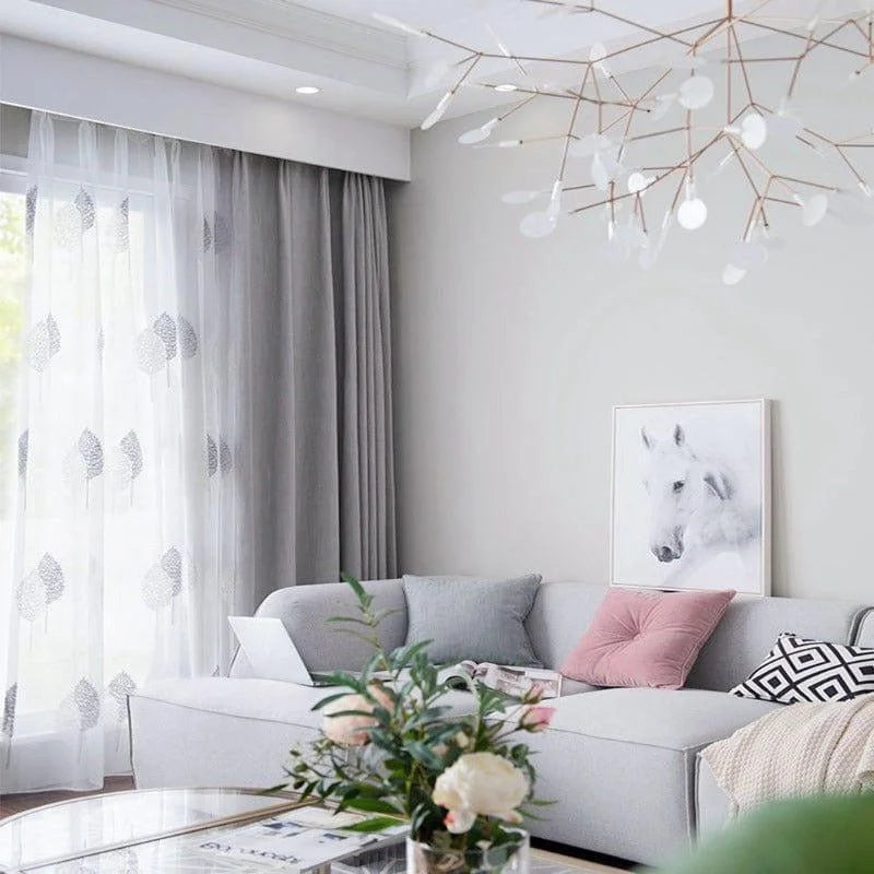 Salon moderne avec décoration élégante, composé d'un canapé angulaire gris clair, agrémenté de coussins variés, dans un intérieur lumineux. Un tableau représentant un cheval blanc est accroché au mur. Un lustre design suspendu au plafond et des rideaux translucides ornent la fenêtre, créant une ambiance chic et confortable idéale pour un espace de vie contemporain.