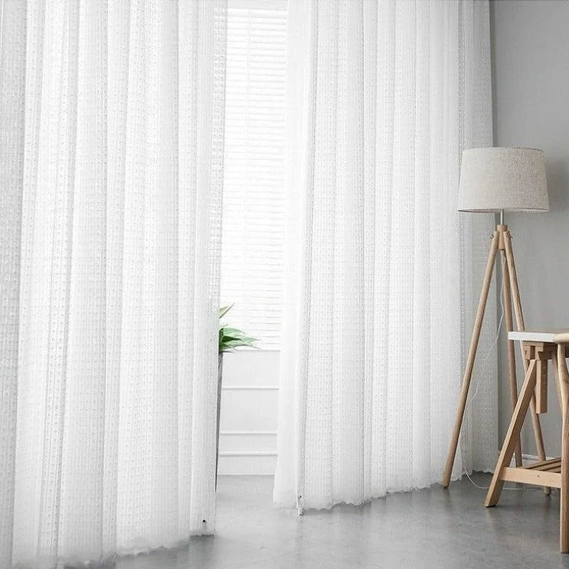 Rideaux blancs transparents suspendus dans un salon moderne, illuminant la pièce avec une lumière douce et naturelle. Ambiance épurée et élégante rehaussée par une lampe sur trépied et un décor minimaliste. Parfaits pour créer une atmosphère accueillante et lumineuse dans tout intérieur.