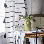 Rideau blanc avec motifs géométriques noirs, suspendu à une fenêtre lumineuse, accompagné d'un vase vert contenant des fleurs violettes, apportant une touche élégante à la décoration intérieure.