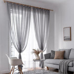 Rideaux gris élégants suspendus dans un salon moderne au design épuré. Ces voilages apportent une touche de sophistication tout en permettant une belle diffusion de la lumière naturelle. Le salon est agrémenté d'un canapé gris confortable et d'une décoration harmonieuse qui inclut un tableau minimaliste et une table basse moderne avec un vase décoratif. Ambiance intérieure chic et contemporaine idéale pour un espace de vie accueillant.
