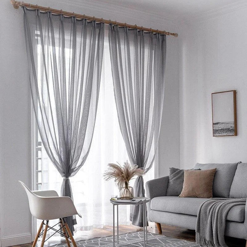 Rideaux gris élégants suspendus dans un salon moderne au design épuré. Ces voilages apportent une touche de sophistication tout en permettant une belle diffusion de la lumière naturelle. Le salon est agrémenté d'un canapé gris confortable et d'une décoration harmonieuse qui inclut un tableau minimaliste et une table basse moderne avec un vase décoratif. Ambiance intérieure chic et contemporaine idéale pour un espace de vie accueillant.