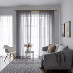 Salon moderne avec rideaux lumineux, canapé gris élégant, tapis à motifs géométriques et fauteuil design. Ambiance cosy et minimaliste pour un espace de vie contemporain.
