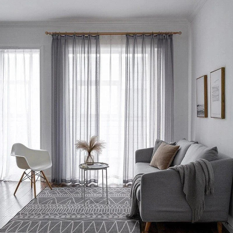 Salon moderne avec rideaux lumineux, canapé gris élégant, tapis à motifs géométriques et fauteuil design. Ambiance cosy et minimaliste pour un espace de vie contemporain.