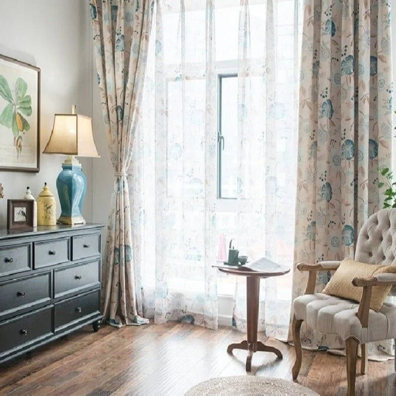 Rideaux élégants avec motifs floraux bleus et beiges, apportant une touche de fraîcheur et de sophistication à un salon lumineux. Les rideaux encadrent une grande fenêtre, permettant une abondance de lumière naturelle. Le décor harmonieux inclut un fauteuil confortable, une table d'appoint en bois et une commode noire avec une lampe et des objets de décoration raffinés, créant une ambiance chaleureuse et accueillante.