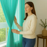 Jeune femme blanche touchant un voilage bleu turquoise dans un intérieur lumineux et épuré, près d'une fenêtre ouverte sur la nature.