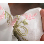 Détail d'un motif floral délicatement brodé avec des fils verts et roses sur un tissu blanc, créant un design élégant et raffiné pour une décoration intérieure ou un accessoire de mode unique.