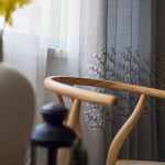 Chaise design en bois avec accoudoirs élégants située près d'une fenêtre ornée de rideaux à motifs floraux, apportant une ambiance chaleureuse et naturelle à l'intérieur. Parfait pour compléter une décoration moderne et raffinée. Idéal dans un salon ou un espace détente.