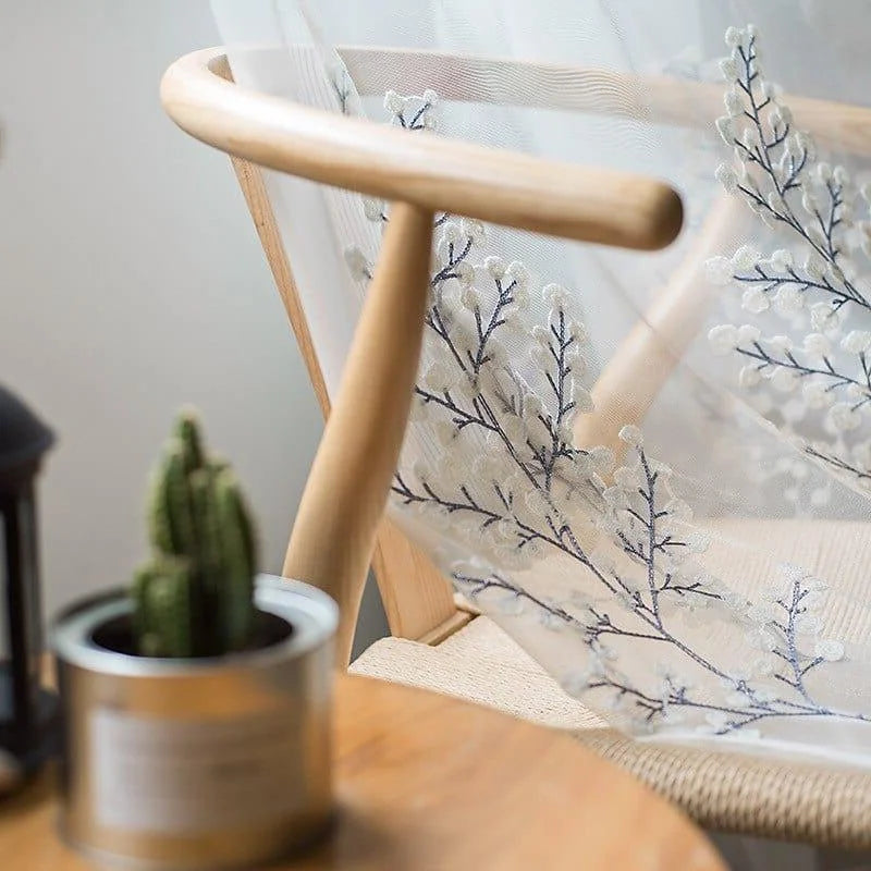 Voilage décoratif avec motif floral délicat, idéal pour un salon moderne. Parfait pour embellir les fenêtres tout en apportant une touche élégante et naturelle. Intégré dans un intérieur chaleureux, il se marie bien avec des éléments en bois et des plantes vertes. Convient à différents styles de décoration.
