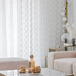 Salon élégant avec rideaux blancs à motifs géométriques, créant une ambiance lumineuse. Canapé moderne couleur beige, table basse en marbre avec objets décoratifs dorés. Parfaite harmonie de design intérieur contemporain.