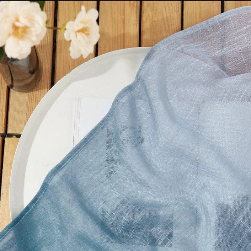 Image d'un élégant tissu bleu pastel posé sur une table en bois, à côté d'un vase contenant des fleurs blanches. Le design présente un motif discret et raffiné, apportant une touche de sophistication et de douceur à la décoration intérieure.