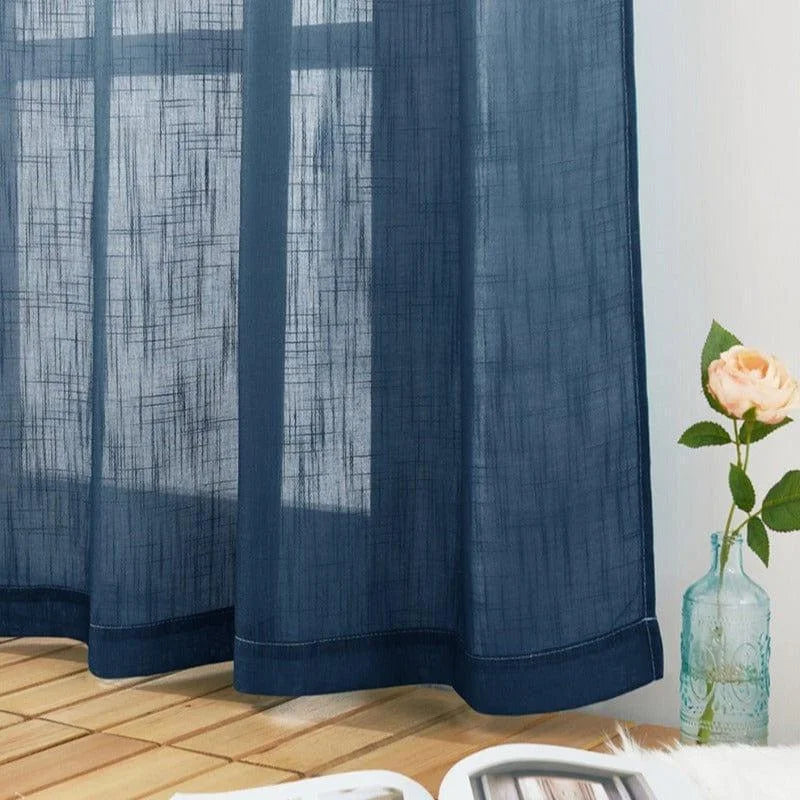 Rideau bleu translucide suspendu à une fenêtre, ajoutant une touche élégante à l'intérieur d'une pièce lumineuse. Une rose en pot et des revues posées sur le sol en bois complètent la décoration chaleureuse et moderne.