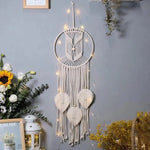 Décoration murale bohème attrape-rêves avec motifs de feuilles, illuminée par des guirlandes lumineuses, sur fond de mur gris, entourée de plantes décoratives et fleurs colorées. Parfait pour une ambiance intérieure chaleureuse et apaisante.