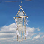 Décoration murale bohème suspendue, composée d'un cercle central avec des plumes blanches et des ornements festonnés, se détachant sur un ciel bleu. Idéale pour une ambiance relaxante et artisanale.