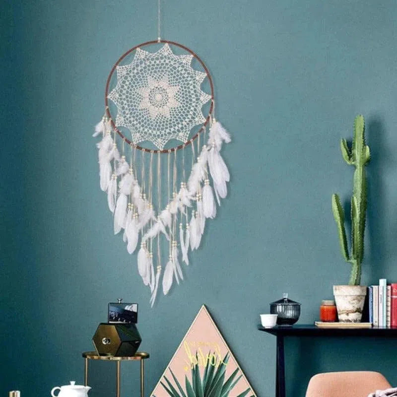 Attrape-rêves décoratif avec un design délicat suspendu sur un mur bleu. Ses plumes blanches et ses détails élégants apportent une touche bohème à l'intérieur. À côté, un cactus et une petite étagère ajoutent à l'atmosphère apaisante et moderne de la pièce. Idéal pour embellir un espace de vie avec style et charme.
