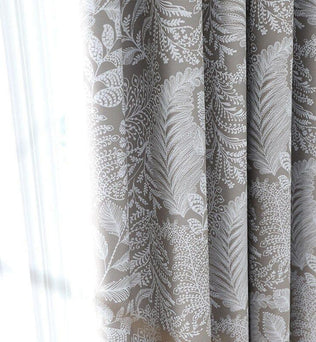 Gros plan sur un rideau américain taupe à motifs blancs végétaux, filtrant la lumière naturelle d'une fenêtre.
