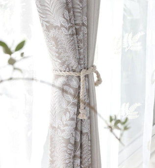 Rideau américain taupe à motifs blancs, attaché avec un cordon torsadé en corde naturelle devant une fenêtre lumineuse.