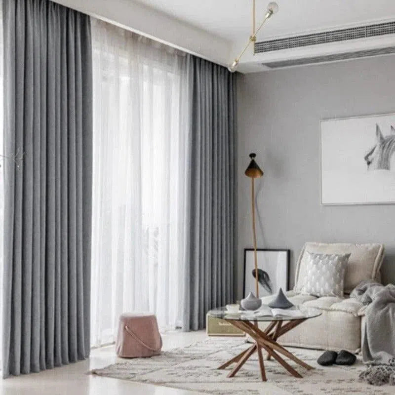 Salon moderne avec de longs rideaux gris élégants, idéal pour une décoration intérieure contemporaine. Les rideaux entourent une grande baie vitrée, apportant luminosité et élégance. Le fauteuil confortable et la table basse en bois ajoutent une touche chaleureuse à l'ensemble. Parfait pour créer une ambiance cosy et accueillante dans votre maison.