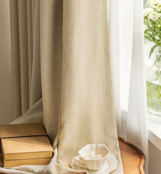 Rideaux élégants couleur beige avec motif chevrons pour salon moderne. Accessoires décoratifs assortis, incluant un vase avec fleurs blanches, posés sur une table en bois éclairée par la lumière naturelle. Ambiance chaleureuse et sophistiquée pour une décoration harmonieuse.