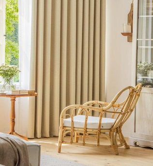 Fauteuil en rotin au design élégant dans un salon lumineux avec des rideaux beiges, une table d'appoint avec des livres et un vase de fleurs, créant une atmosphère chaleureuse et accueillante.