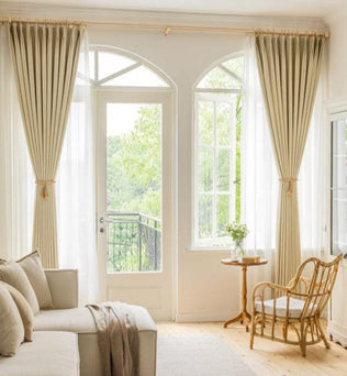 Salon élégant avec rideaux beiges, canapé confortable et chaise en osier, offrant une ambiance lumineuse et chaleureuse. Baie vitrée donnant sur un balcon avec vue sur la nature verdoyante. Aménagement idéal pour une décoration intérieure moderne et sereine.