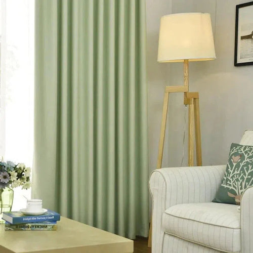 Salon moderne avec rideaux verts, lampe sur pied en bois et fauteuil blanc à rayures fines. Décoration intérieure élégante avec des coussins décoratifs sur le canapé, une table basse avec livres et fleurs en vase. Atmosphère accueillante et lumineuse.