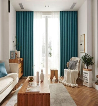 Salon moderne et élégant avec un canapé beige et un coussin bleu, rideaux turquoise, table basse en bois et fauteuil enveloppé d’un plaid. Décoration intérieure lumineuse et soignée, plancher en bois, plantes vertes et accents déco harmonieux. Large fenêtre laissant entrer une lumière naturelle abondante, créant une ambiance chaleureuse et accueillante. Idéal pour inspiration décoration ou aménagement intérieur.