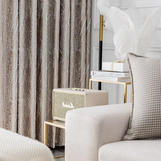 Image d'un salon élégant avec rideaux texturés marron, un canapé confortable et un coussin à motifs. Une lampe stylisée et des livres sur une table d'appoint dorée ajoutent une touche de sophistication. Une enceinte vintage complète l'ambiance chic et moderne.