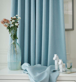 Rideaux élégants de couleur bleu clair dans un intérieur moderne, accompagnés d'un vase décoratif avec des fleurs fraîches sur une table blanche, parfaits pour ajouter une touche de sérénité à votre salon ou chambre.