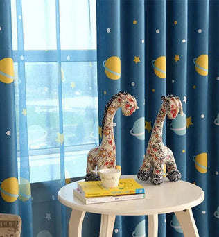 Rideau enfant motif espace bleu avec planètes jaunes, parfait pour créer une ambiance cosmique dans la chambre. Décoration originale avec girafes en peluche sur une table ronde blanche, ajoutant une touche ludique et chaleureuse. Idéal pour stimuler l'imagination des enfants tout en embellissant leur espace de vie.