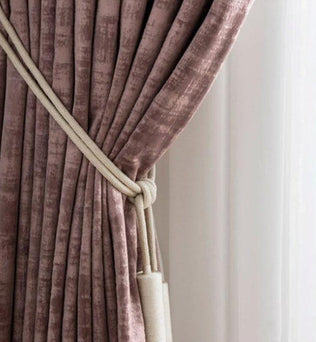 Rideau élégant de couleur rose poudre, replié avec un embrasse de cordon beige. Ce rideau habille une fenêtre en ajoutant une touche de sophistication et de style chaleureux à l'intérieur de la pièce. Idéal pour créer une ambiance cosy et chic dans le salon ou la chambre.