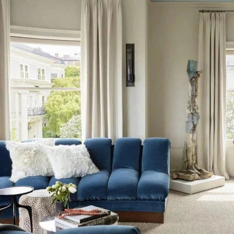Salon contemporain avec canapé bleu, coussins blancs et table basse moderne. Grandes fenêtres avec rideaux blancs offrant une vue sur l'extérieur verdoyant. Ambiance lumineuse et élégante pour un intérieur design.