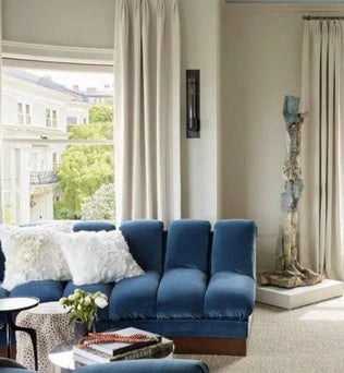 Salon contemporain avec canapé bleu, coussins blancs et table basse moderne. Grandes fenêtres avec rideaux blancs offrant une vue sur l'extérieur verdoyant. Ambiance lumineuse et élégante pour un intérieur design.