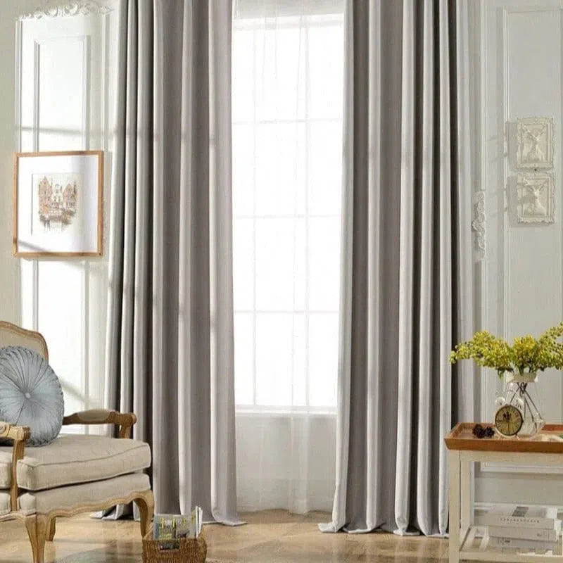 Rideaux élégants de couleur grise suspendus dans un salon lumineux. Ambiance moderne et sophistiquée avec des meubles classiques, un fauteuil confortable et une table d'appoint décorée de fleurs jaunes. Idéal pour sublimer la décoration intérieure et apporter une touche de raffinement.