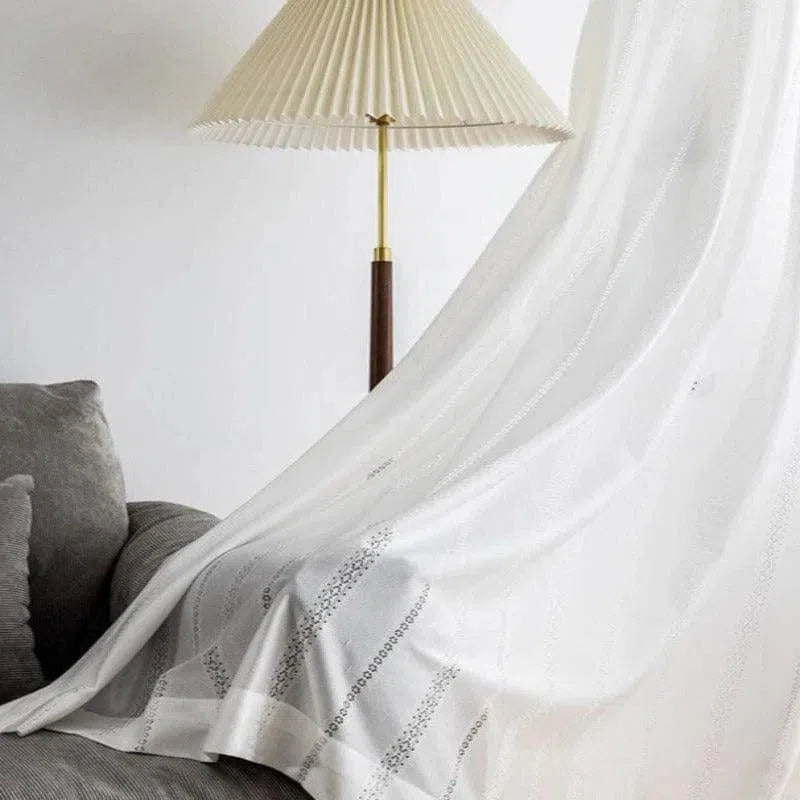 Rideau blanc élégant avec motifs délicats, installé près d'un canapé gris et d'une lampe à pied, apportant une touche moderne et lumineuse à la décoration intérieure.