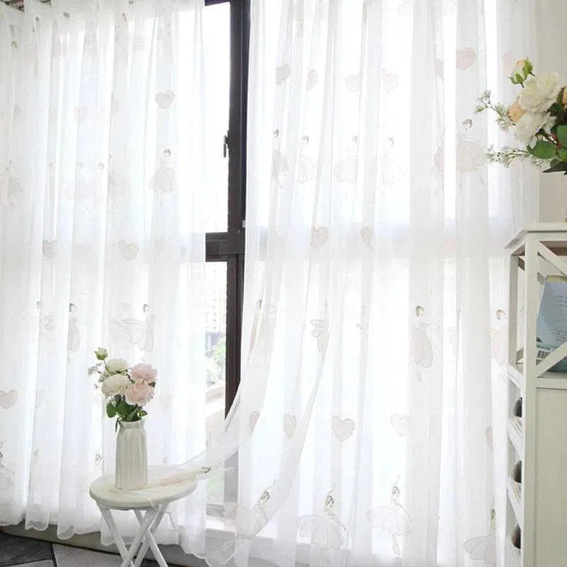 Rideau blanc avec motifs de cœurs suspendu devant une fenêtre, offrant une ambiance lumineuse et élégante à la pièce. Une table blanche avec un vase de fleurs roses apporte une touche de fraîcheur. Idéal pour une décoration intérieure moderne et épurée.