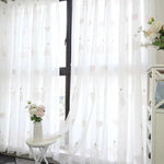 Rideau blanc avec motifs de cœurs suspendu devant une fenêtre, offrant une ambiance lumineuse et élégante à la pièce. Une table blanche avec un vase de fleurs roses apporte une touche de fraîcheur. Idéal pour une décoration intérieure moderne et épurée.