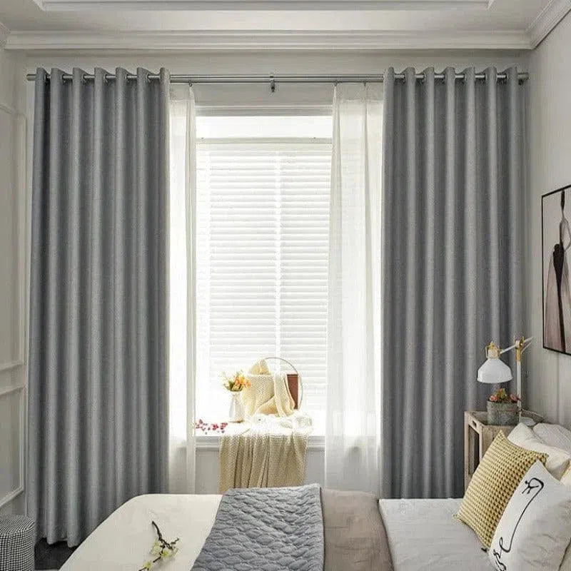 Chambre moderne et élégante avec rideaux opaques gris encadrant une fenêtre lumineuse; lit confortable avec coussins jaunes et gris, créant une ambiance douce et accueillante. Décoration intérieure épurée parfaite pour un espace de détente, combinant style et fonctionnalité.