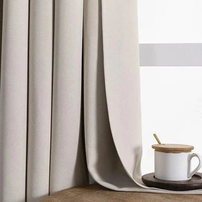 Rideau beige occultant élégant en tissu épais, idéal pour la décoration intérieure moderne. Offre intimité et contrôle de la lumière, parfait pour chambre ou salon. Assorti à une tasse en céramique blanche sur une table en bois clair. Acheter des rideaux de qualité pour un intérieur contemporain et chaleureux.