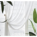 Rideau blanc léger drapé devant une fenêtre avec des stores, entouré de plantes vertes. Crée une ambiance douce et lumineuse, idéal pour un salon moderne. Décoration intérieure élégante et naturelle.