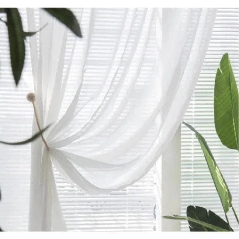 Rideau blanc léger drapé devant une fenêtre avec des stores, entouré de plantes vertes. Crée une ambiance douce et lumineuse, idéal pour un salon moderne. Décoration intérieure élégante et naturelle.