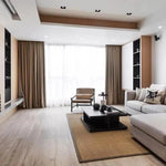 Salon contemporain avec canapé d'angle gris, fauteuil élégant, table basse noire et décorations minimalistes. Grandes fenêtres avec rideaux marron, parquet clair et ambiance lumineuse pour un espace de vie moderne et accueillant.