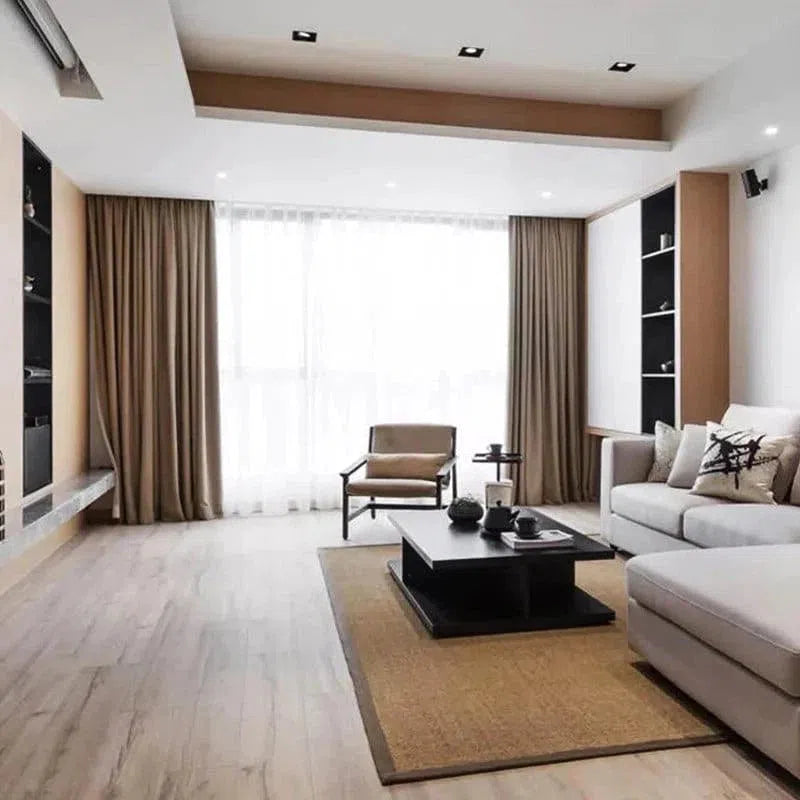 Salon contemporain avec canapé d'angle gris, fauteuil élégant, table basse noire et décorations minimalistes. Grandes fenêtres avec rideaux marron, parquet clair et ambiance lumineuse pour un espace de vie moderne et accueillant.
